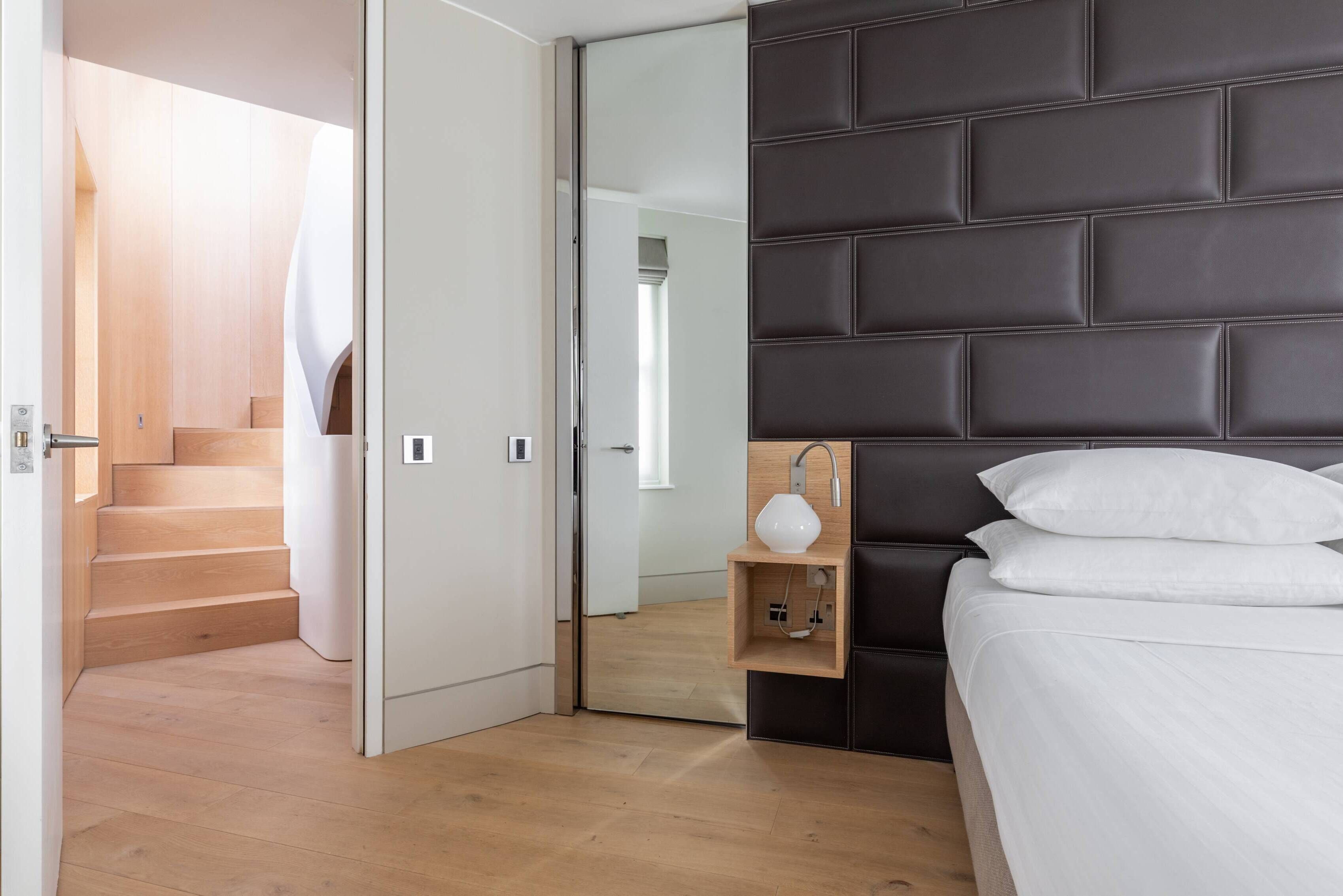 A modern bedroom with a wooden floor, a dark headboard, a bedside table with a lamp, and a bed with white bedding.