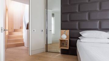 A modern bedroom with a wooden floor, a dark headboard, a bedside table with a lamp, and a bed with white bedding.