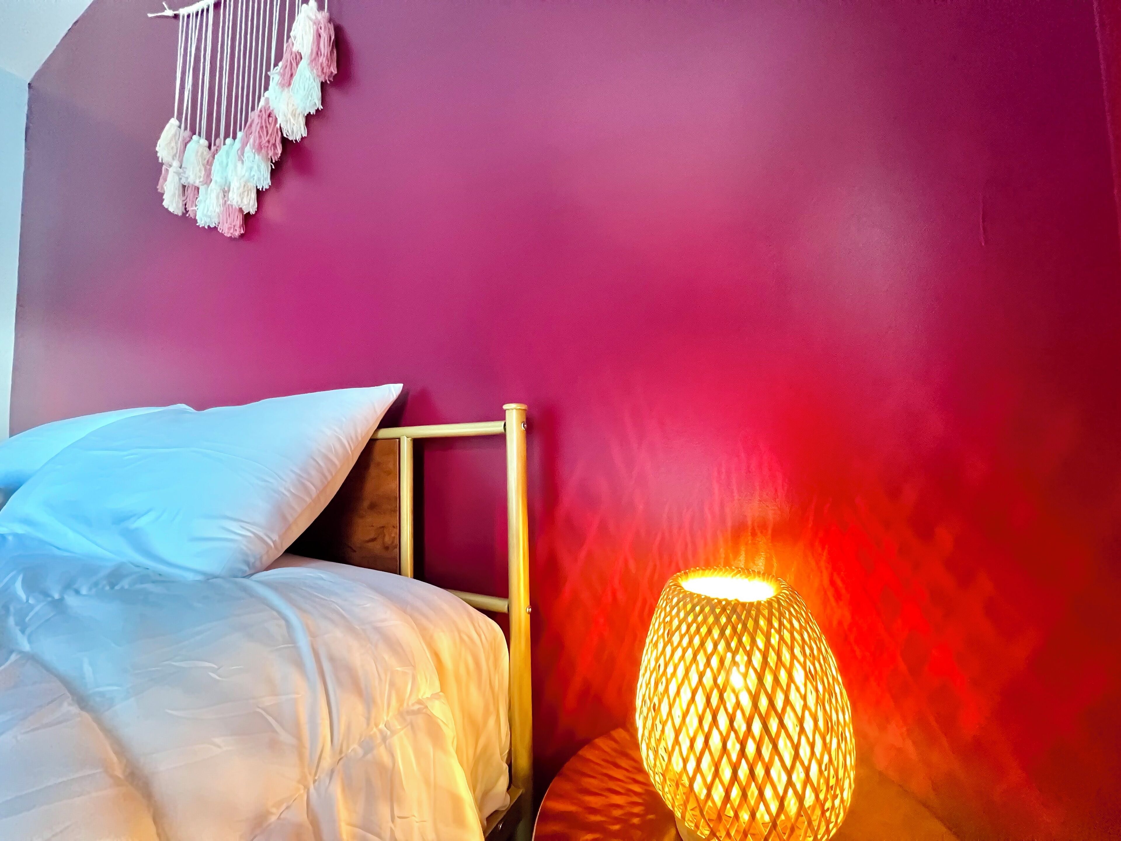 Una cama con ropa de cama blanca, una lámpara de noche y un tapiz de macramé.