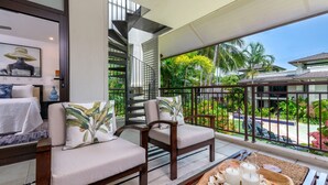 Een balkon met een bank en stoelen, een houten tafel met kaarsen, en uitzicht op een zwembad en tropische planten.