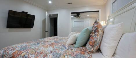 A bedroom with a bed, a ceiling fan, a TV mounted on the wall, and a door leading to another room.