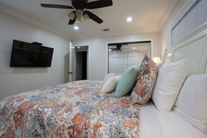 A bedroom with a bed, a ceiling fan, a TV mounted on the wall, and a door leading to another room.