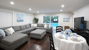 A modern living room with a gray sectional sofa, a dining table set for four, and a large flat-screen TV. - Sweet 5 Bedroom, 4 Bath Pool Home 1/2 Mile Walk To Disney (Anaheim)