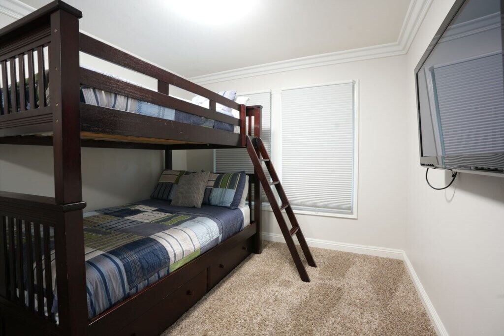 A bunk bed with a ladder, a window with blinds, and a television on a stand.