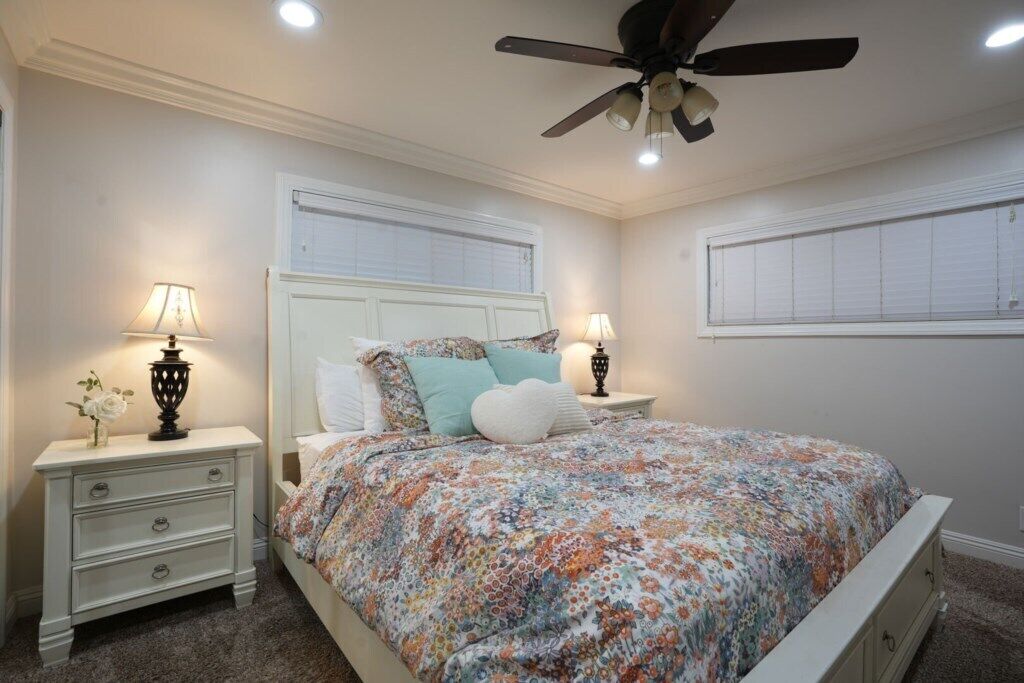 A bedroom with a bed, two bedside tables with lamps, a ceiling fan, and a window with blinds.