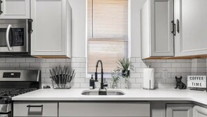 A modern kitchen with white cabinetry, a stainless steel range hood, a built-in microwave, a sink with a black faucet, and a window with blinds. - Cozy & Stylish/Pool Table & Fun

 (Camden)