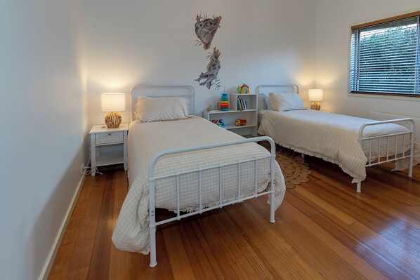 A bedroom with two beds, a nightstand, a bookshelf, and a window with blinds. - Absolute lake frontage property located in the heart of beautiful Mallacoota. (Mallacoota)