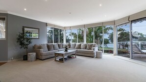 A spacious living room with a large grey sofa, a coffee table, and a view of the outdoors through large windows. - Absolute lake frontage property located in the heart of beautiful Mallacoota.  (Mallacoota)