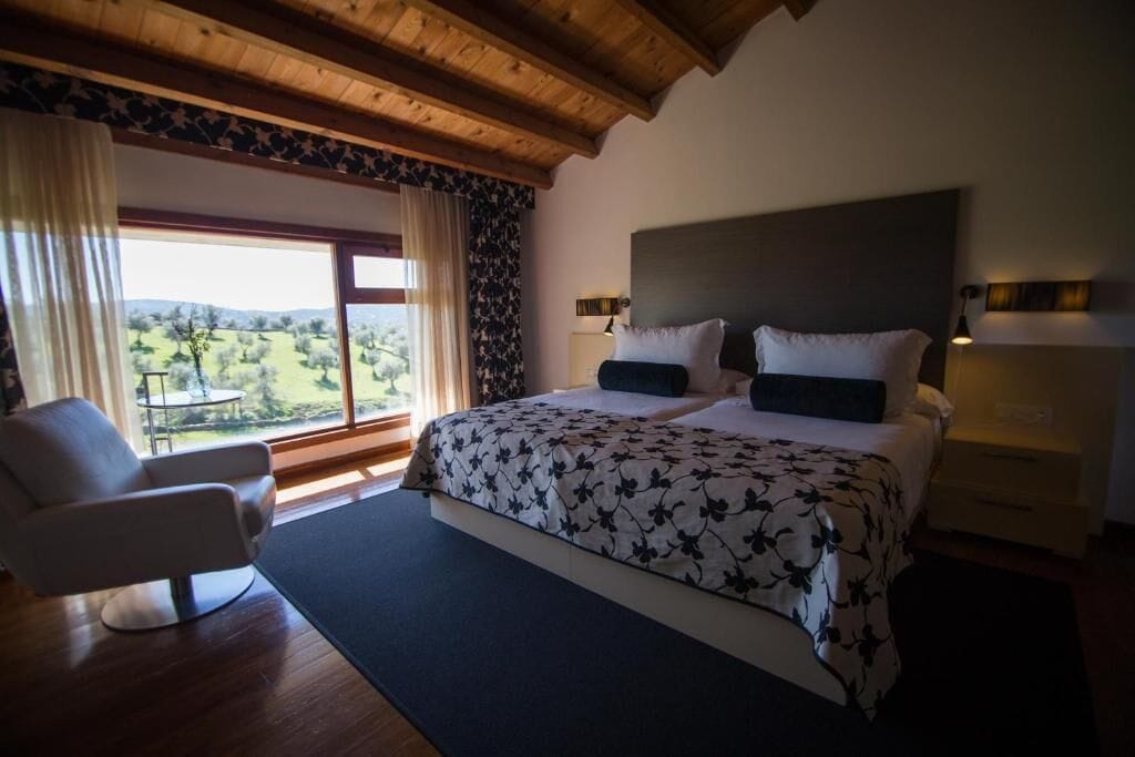 A bedroom with a large bed, a chair, a nightstand, and a view of the outdoors.