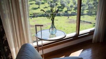 Ein Zimmer mit Blick auf eine grüne Landschaft durch ein Fenster, ein weißer Stuhl und ein Glastisch mit einer Vase.