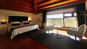 Une chambre à coucher avec un grand lit, deux fauteuils blancs, une petite table et une grande fenêtre donnant sur la campagne.
