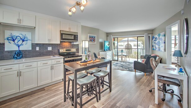 A modern kitchen with white cabinets, a central island with stools, a microwave, and a stove. There is a dining area with a table and chairs, and a large window offering a view of the outdoors. - Beautiful one bedroom condo in Marco Island with so much to offer!  (Marco Island)
