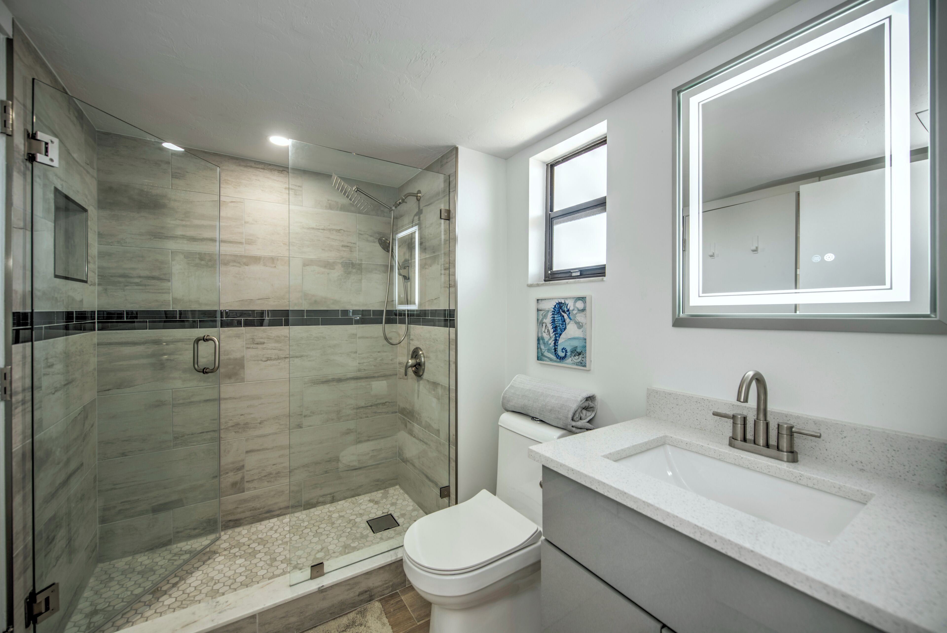 Une salle de bain moderne avec une douche à parois de verre, des toilettes blanches, un lavabo avec un miroir et une fenêtre au-dessus du lavabo.