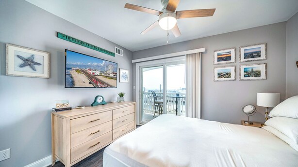 Habitación de hotel con cama, cómoda, ventilador de techo, televisión y balcón con vista a la playa y una noria.