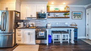 Una cocina moderna con armarios blancos, electrodomésticos de acero inoxidable, microondas y tostadora. Hay estantes de madera sobre la encimera con adornos y una foto enmarcada.