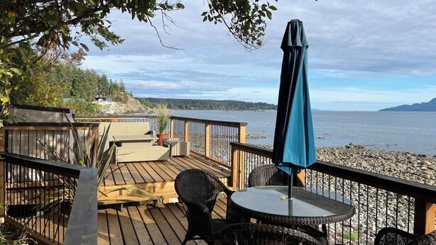 A wooden deck with wicker furniture and a glass table, overlooking a rocky shoreline and a body of water. - Malaspina Strait Tiny Home with easy access to town and trails for hiking/biking (Powell River)