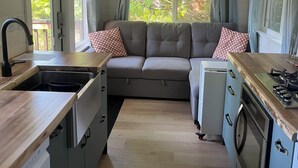 A compact kitchen with a built-in oven, a sink, and a dining area with a grey sofa and patterned cushions. - Malaspina Strait Tiny Home with easy access to town and trails for hiking/biking (Powell River)