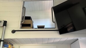 A room with a TV mounted on a white wall, a window with blinds, and a door leading to another room. - Malaspina Strait Tiny Home with easy access to town and trails for hiking/biking (Powell River)