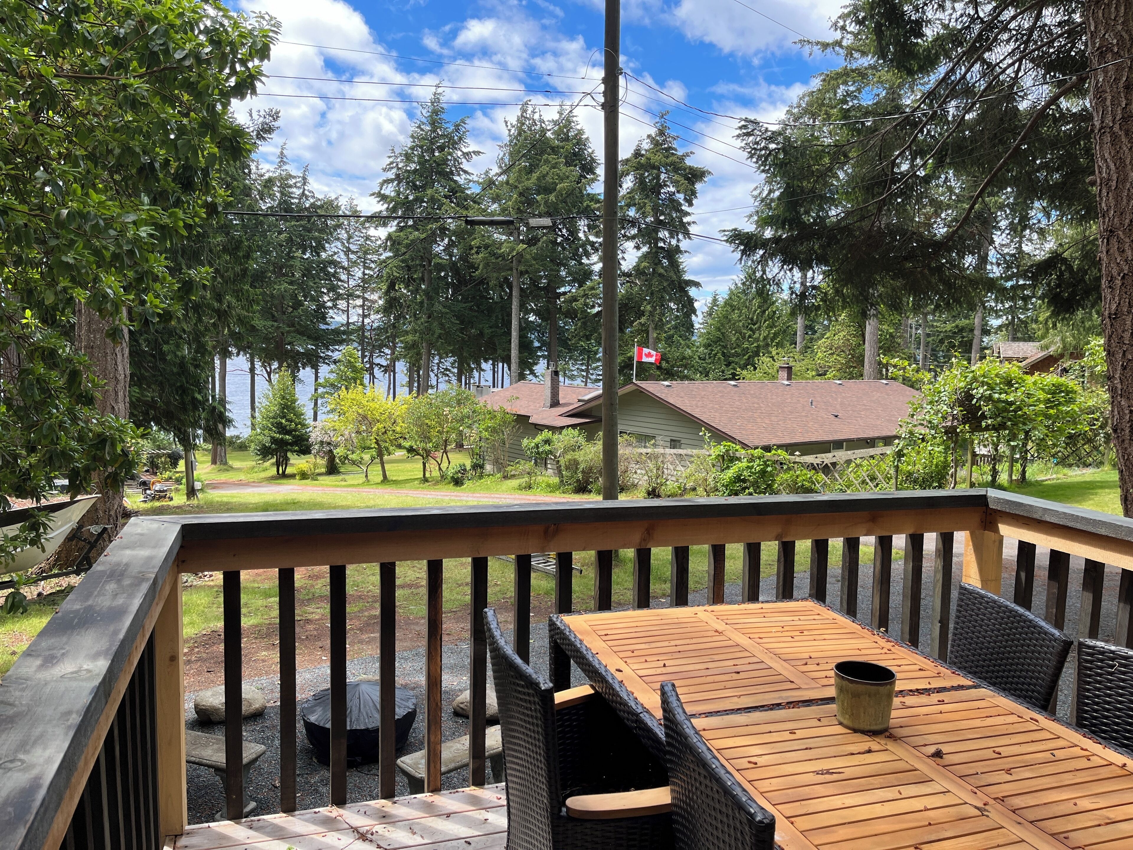 A wooden deck with a table and chairs, overlooking a lake with a house and trees in the background.