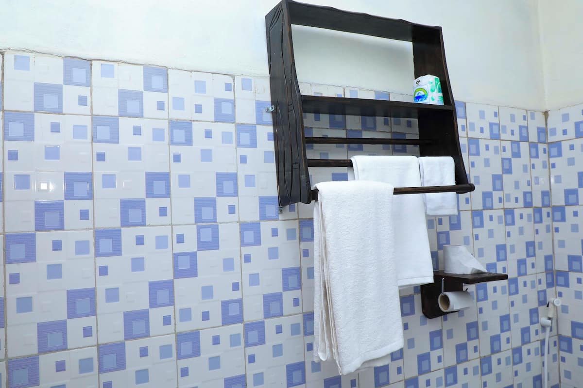 A bathroom with a tiled wall, a towel rack with two white towels, a toilet paper holder, and a shelf with a bottle.