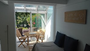 Ein Zimmer mit einem Bett, einem Tisch mit Stühlen, einer Glastür zu einem Außenbereich mit Pflanzen und einem Sessel.
