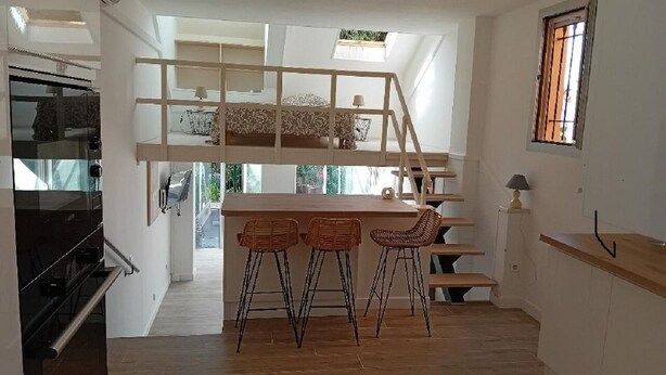 Eine kompakte Küche mit Holzeiland und Barhocker, ein Schlafbereich im Zwischengeschoss mit Etagenbett sowie ein Fenster mit Gitterstäben.