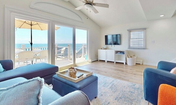 A spacious living room with a large sliding glass door leading to a balcony with a view of the ocean. There are multiple seating areas, a flat-screen TV mounted on the wall, and a ceiling fan. - Charming 3 Bedroom Beach Front Cottage (Milford)