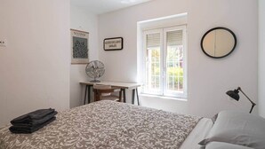 A bedroom with a bed, a desk, a chair, a fan, a window with shutters, and a wall clock.