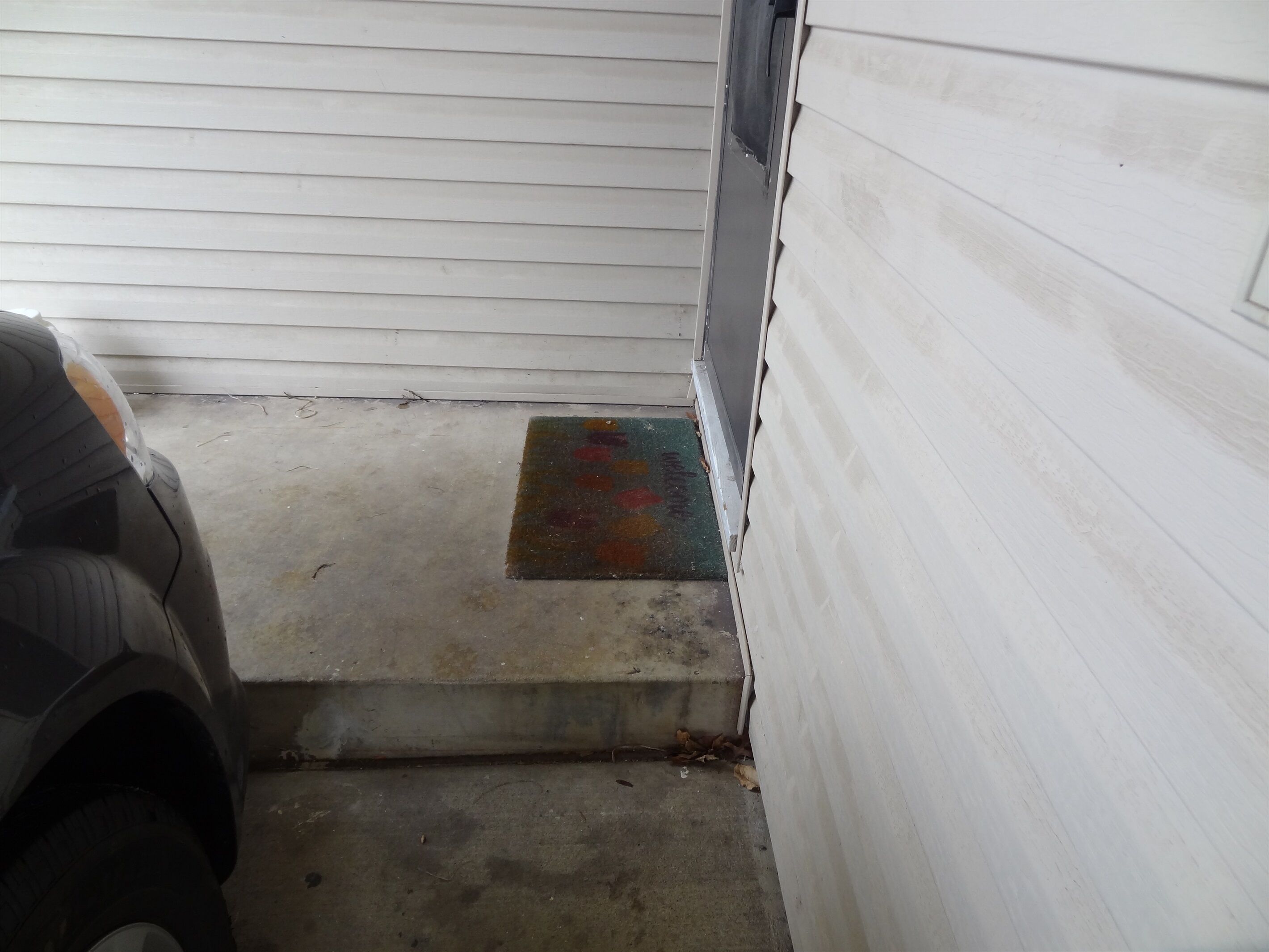A narrow entryway with a concrete floor, a small step leading to a door, and a colorful mat at the door's entrance.