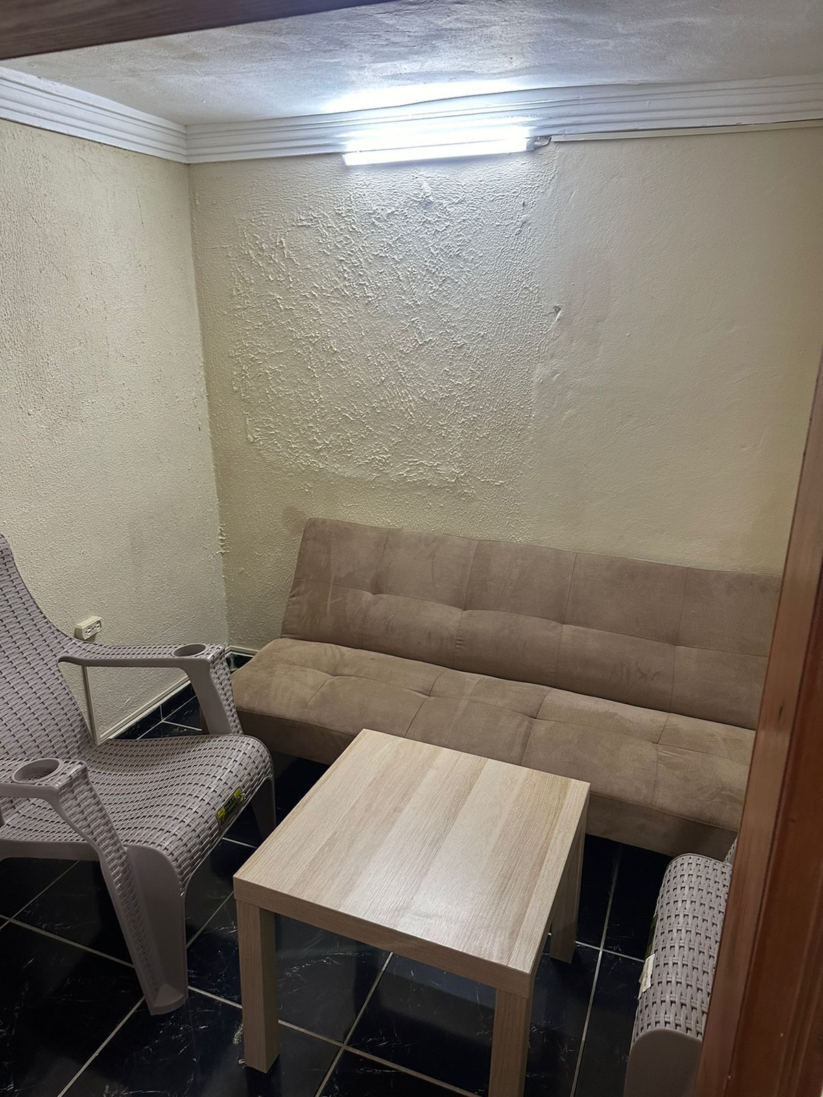 Een kleine, afgesloten kamer met een beige muur met textuur, een bruine bank, een rieten stoel en een houten tafel.
