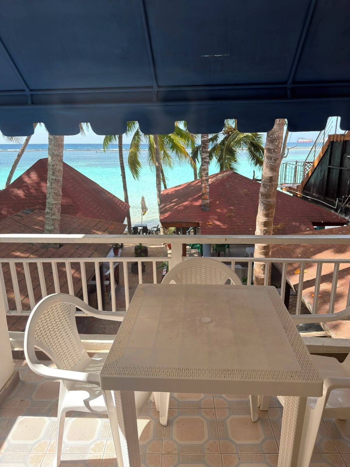 A covered outdoor seating area with a table and chairs, overlooking a beach with palm trees and a building in the background.