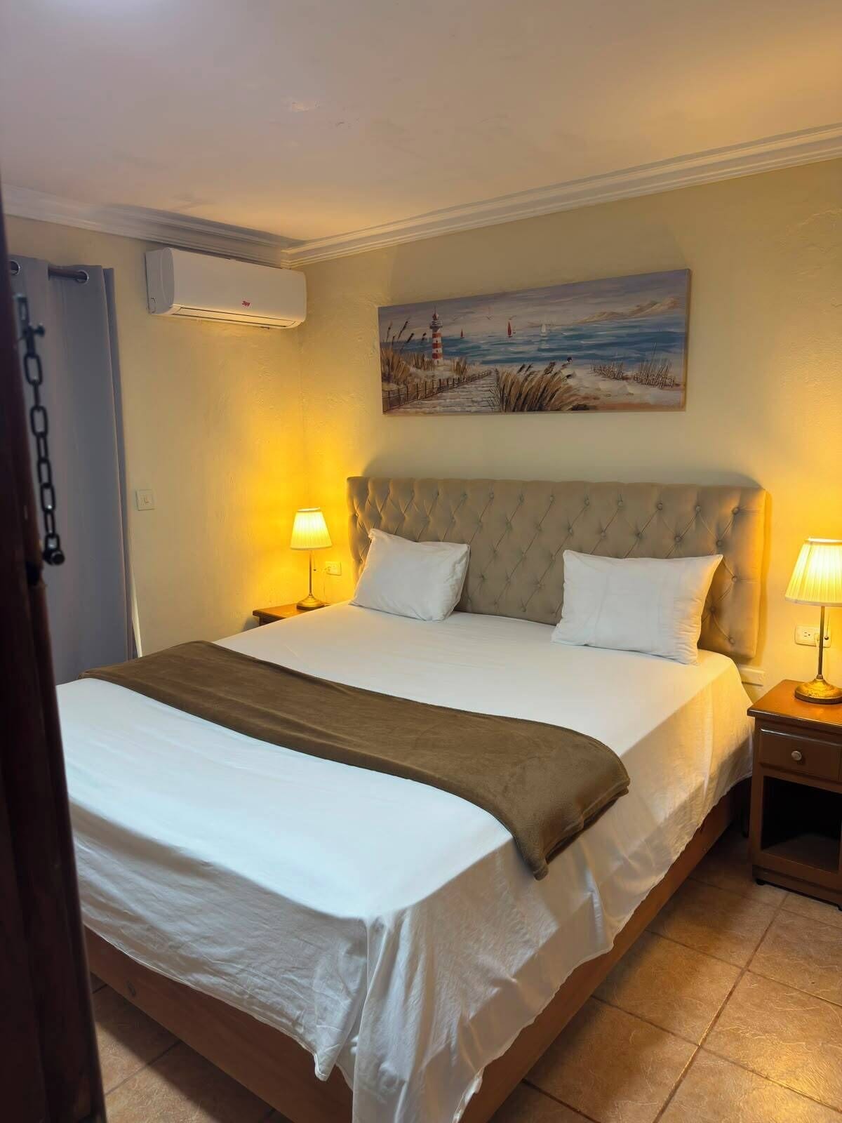 Een hotelkamer met een bed, twee bedlampen, een schilderij aan de muur en een airconditioning.