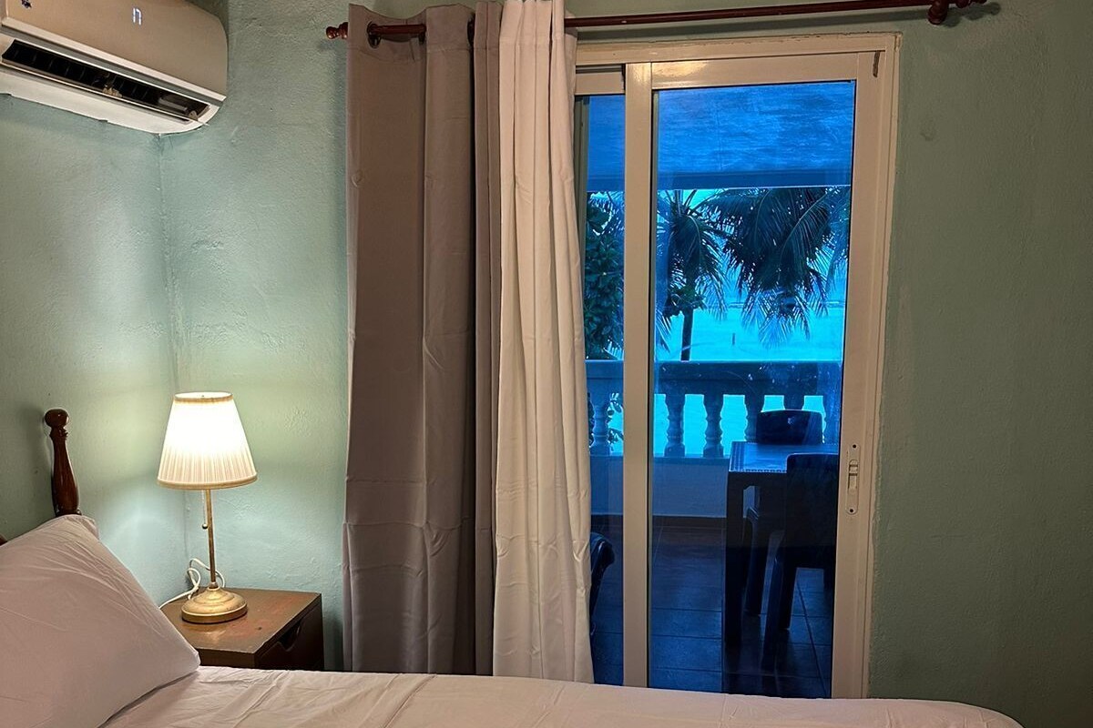 Un dormitorio con una cama, una mesita de noche con lámpara y una puerta corrediza de vidrio que da a un balcón con vistas a palmeras.