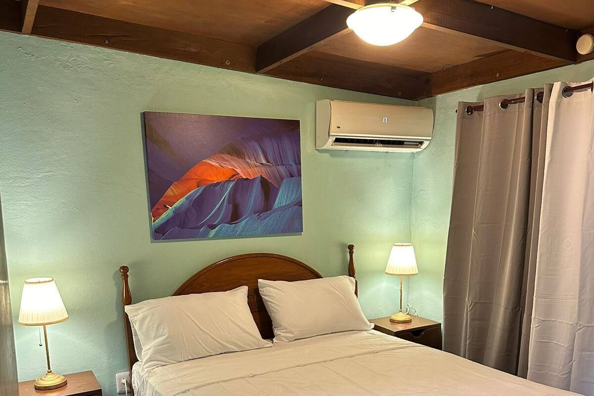 Un dormitorio con una cama, dos mesitas de noche con lámparas, un aire acondicionado instalado en la pared y un cuadro sobre la cama.