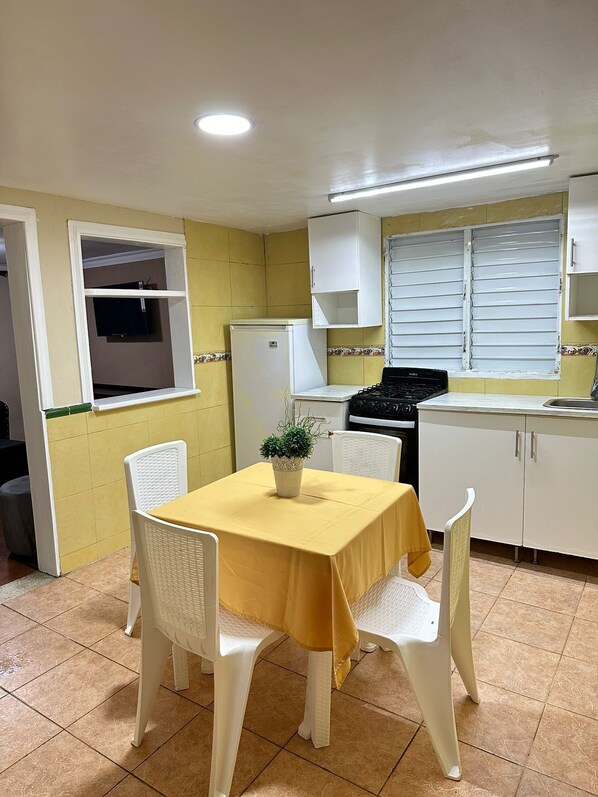 A compact kitchen with a yellow table, white chairs, a refrigerator, a stove, and a window with blinds. - El Cayuco Restaurant apart Hotel (Boca Chica)