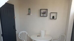Un pequeño patio amurallado con una mesa redonda y cuatro sillas de plástico blancas. Se ve una puerta a la izquierda y una planta pequeña en maceta en la pared.