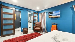 Habitación de hotel con una cama, una alfombra roja estampada, un sillón de cuero marrón, una toalla blanca, una puerta corredera de madera y un cuadro con motivos playeros en la pared.
