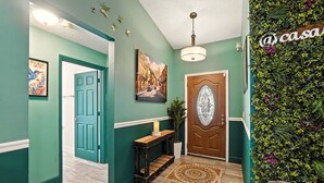 Un pasillo con paredes azul verdoso, una puerta de madera, una pequeña consola con una vela, un tapete estampado y una planta colgante en la pared.