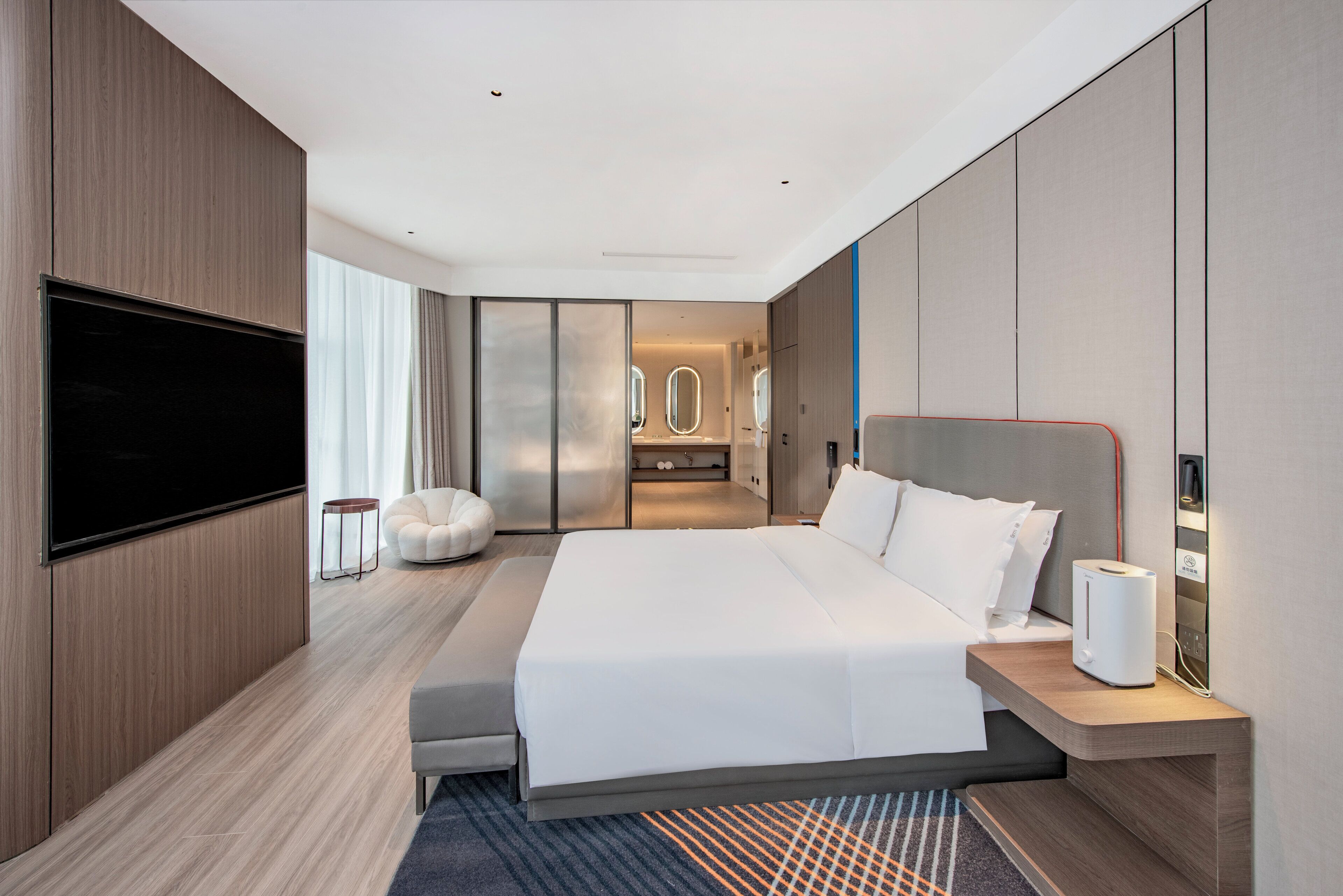 A modern hotel room with a large bed, a flat-screen TV, a seating area with a bean bag, and a view of the bathroom.