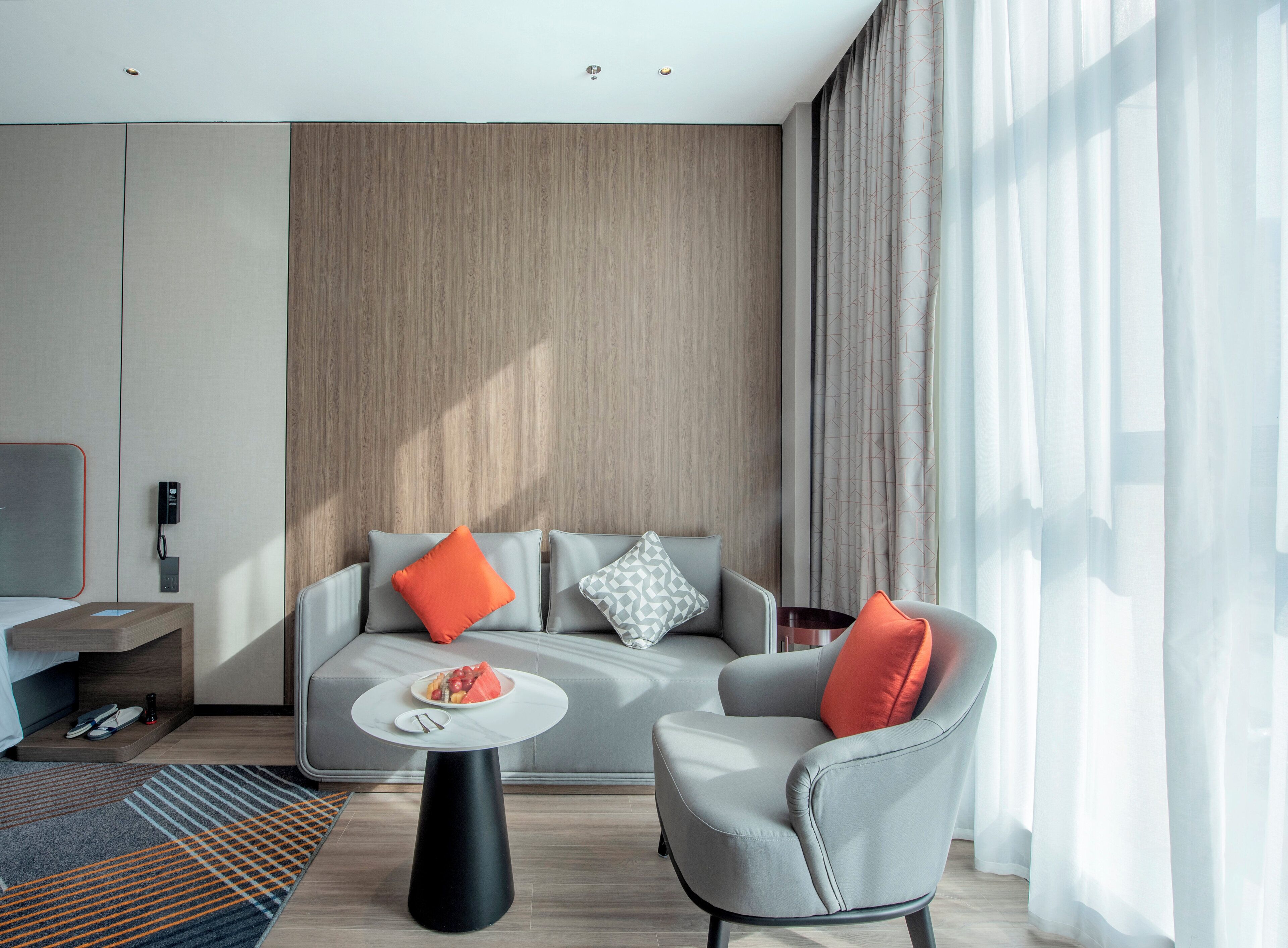 Ein modernes Hotelzimmer mit einer grauen Couch, einem kleinen Sessel, einem runden Couchtisch und einem Flachbildfernseher auf einem Standfuß.