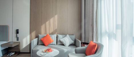 A modern hotel room with a grey sofa, a smaller armchair, a round coffee table, and a flat-screen TV on a stand.