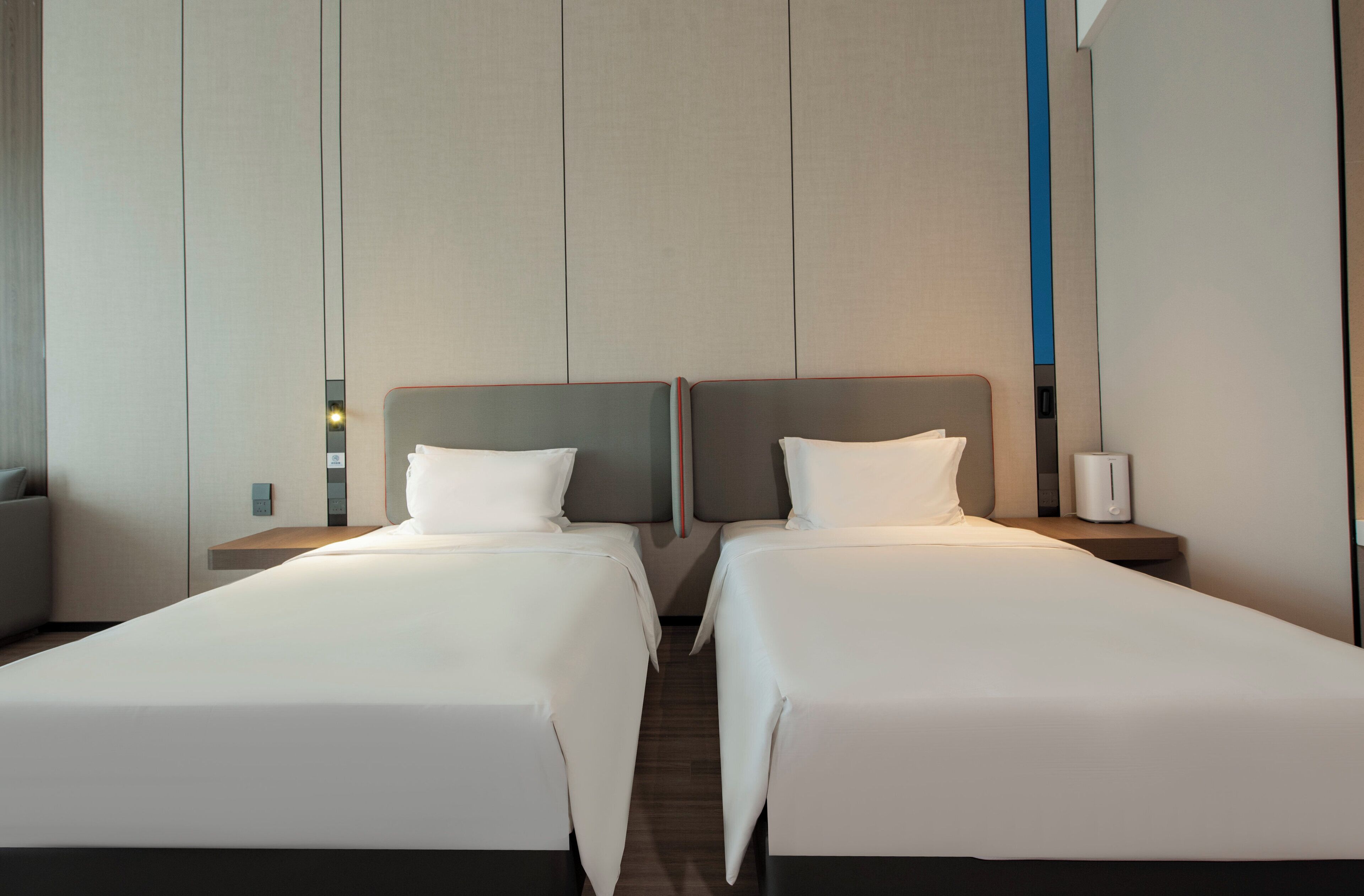 A hotel room with two single beds, a bedside table with a lamp, and a wall-mounted light fixture.