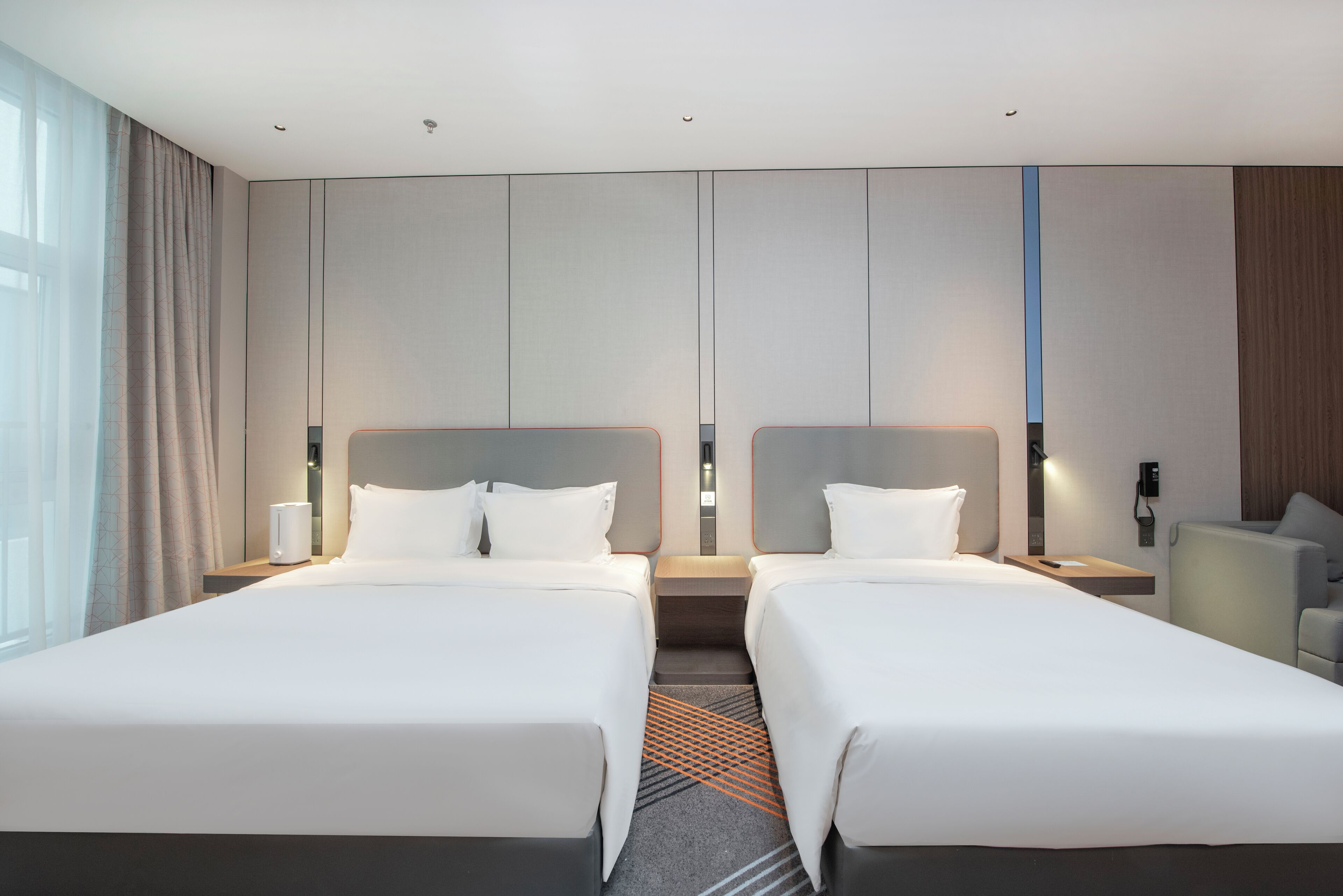 Habitación de hotel con dos camas, una mesita de noche con lámpara y vista a la ciudad.