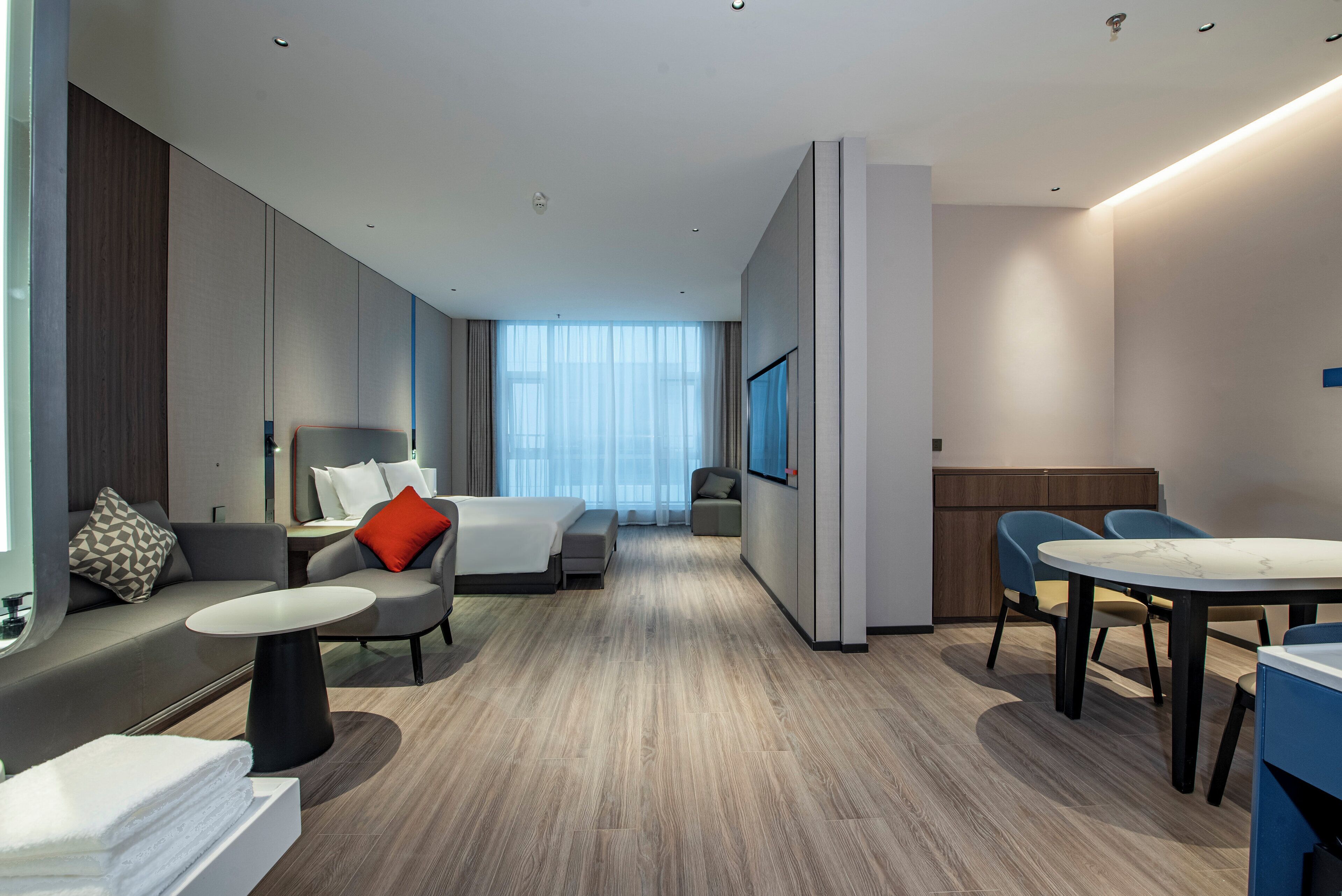 A modern hotel room with a bed, a sofa, a dining table, and a TV.