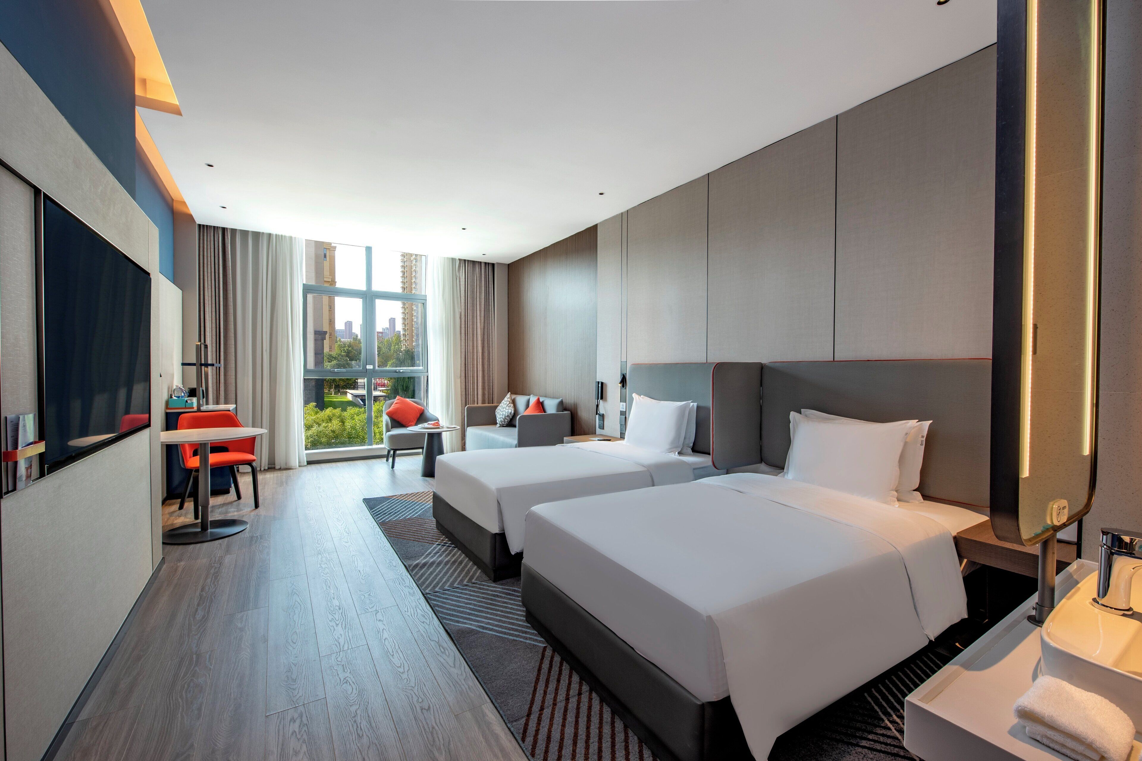 Ein modernes Hotelzimmer mit zwei Betten, einem Schreibtisch und einem großen Fenster mit Blick auf die Stadt.