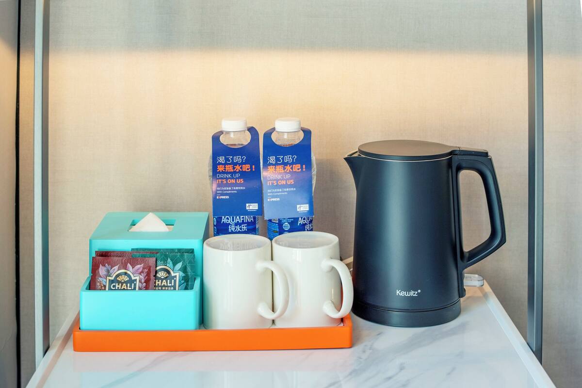 Un vassoio con due tazze bianche, due bottiglie d'acqua Aquafina, una macchina da caffè Keurig e una scatola di fazzoletti con bustine di tè Chali.