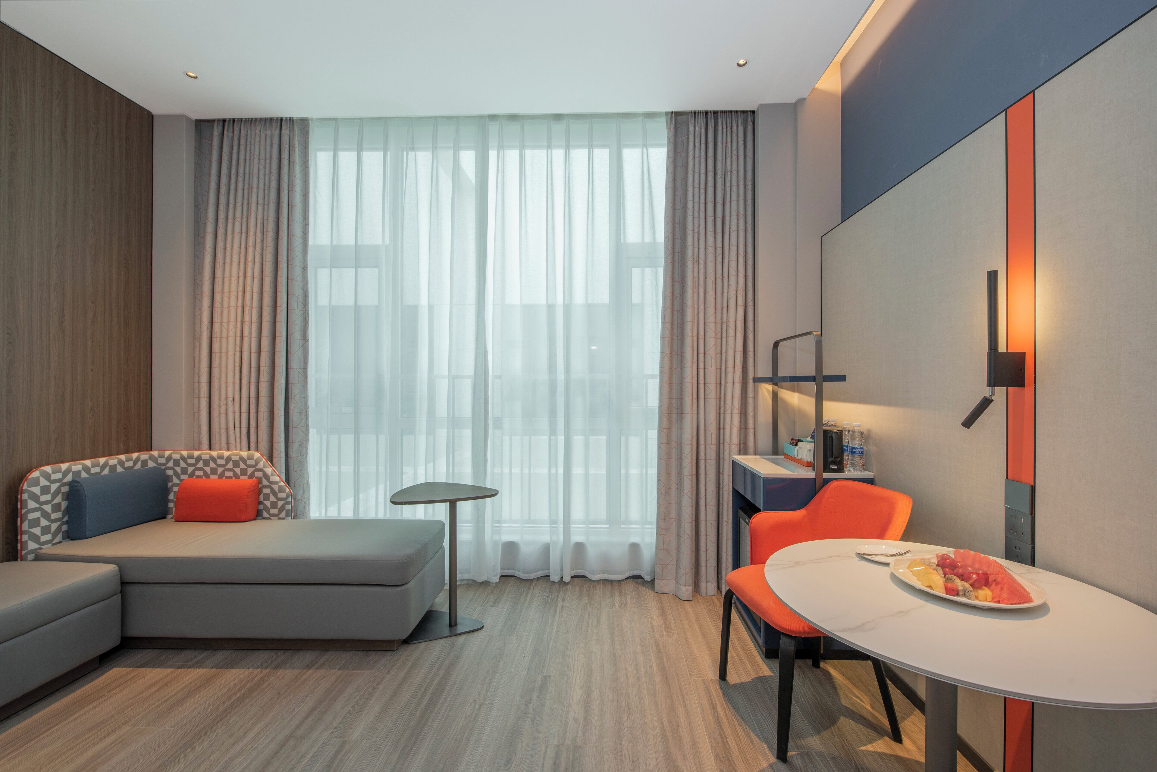 Ein modernes Hotelzimmer mit einer Couch, einem Esstisch mit Obstteller, einem kleinen Tisch und einem Stuhl.