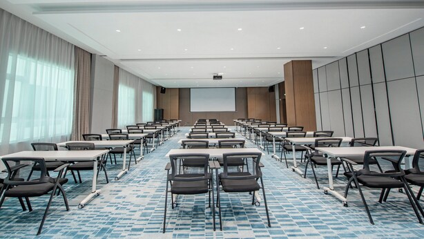 A conference room with rows of tables and chairs, a projector screen, and a large window with curtains. - Holiday Inn Express Yinchuan Lanxi Valley Plaza by IHG (Yinchuan)
