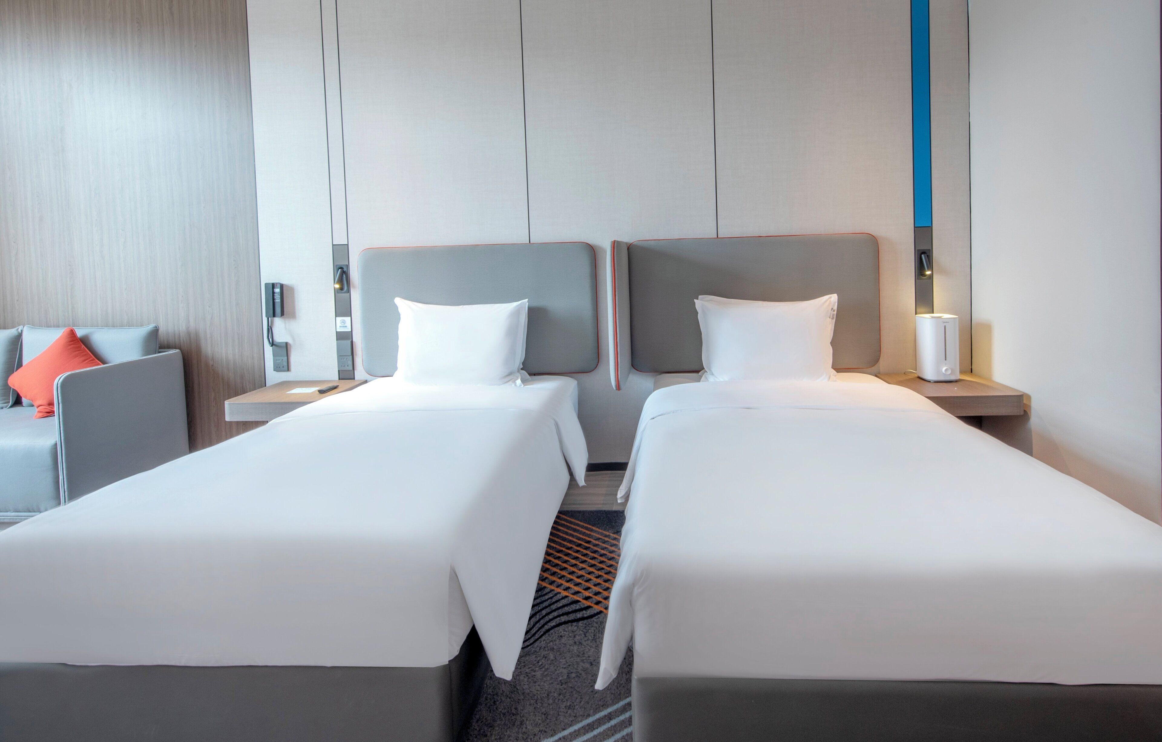 A hotel room with two single beds, a desk, a chair, and a nightstand with a lamp.