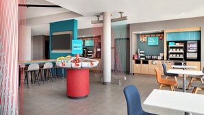 Een modern café met een rode tafel in het midden waarop fruit staat, en een bord met de tekst 'Good round'. Er zijn verschillende zithoeken met stoelen en tafels, en een balie met een koffieapparaat en een menukaart.